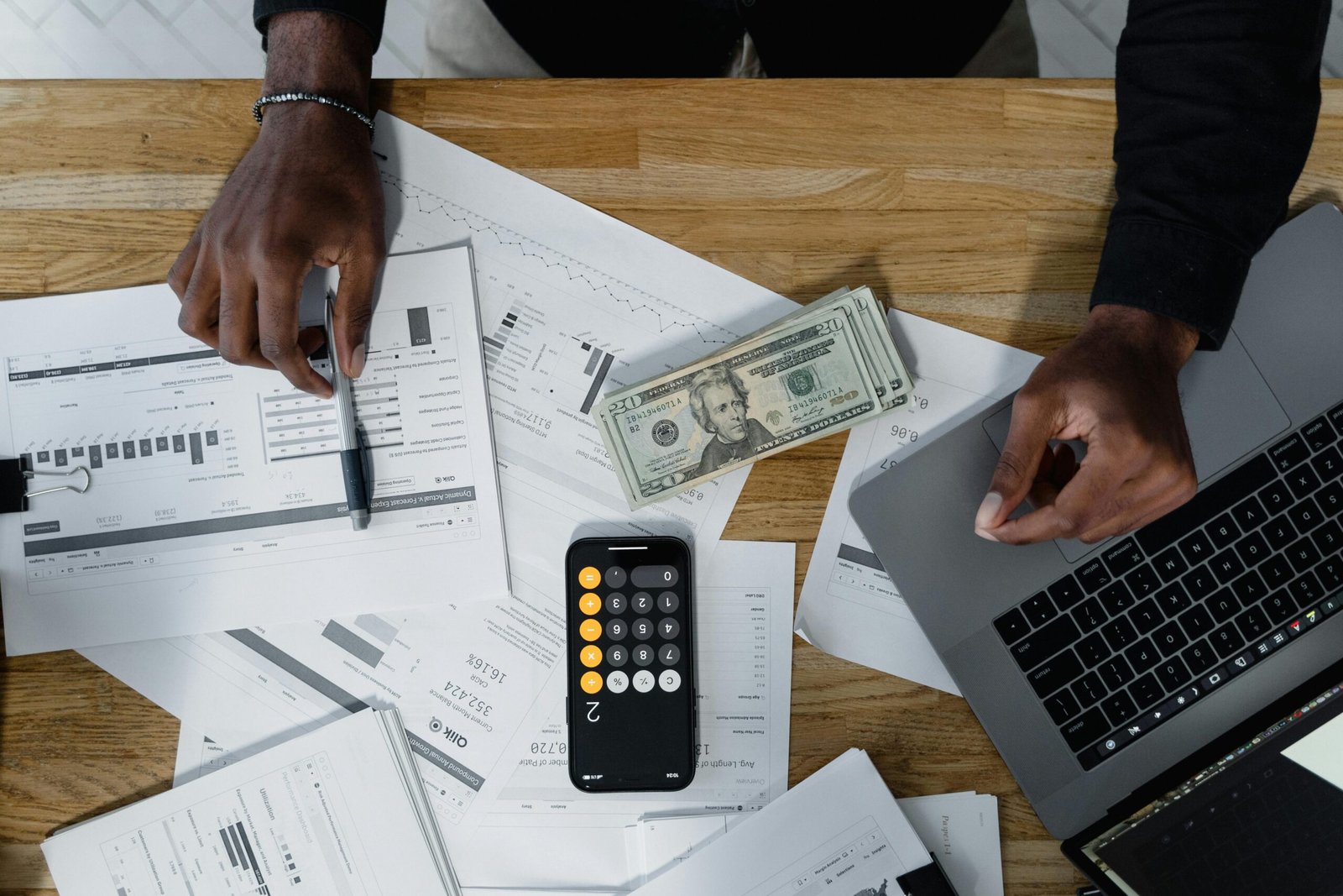 Hands calculate finances with papers, cash, and a laptop on a wooden desk.